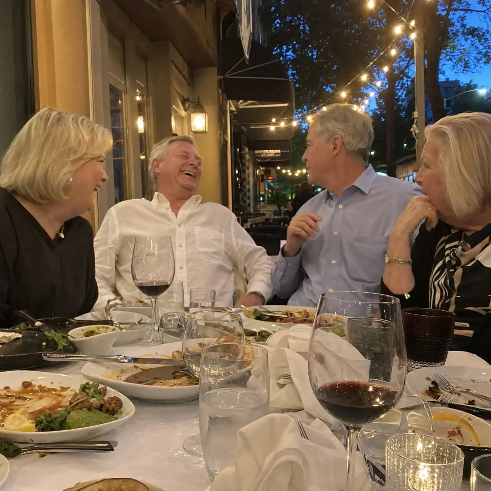 Two couples laughing together over dinner on a restaurant patio at dusk